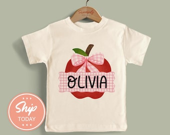 May include: A cream-colored short-sleeve t-shirt featuring a graphic of a red apple with a pink and white gingham bow. A pink and white gingham banner across the center of the apple displays the name OLIVIA in bold black lettering.
