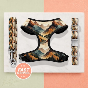 May include: A pet harness, leash, and collar set with a mountain landscape design. The harness and collar have black trim and buckles. The leash has a metal clasp. The set is displayed on a white surface with a peach and green background.