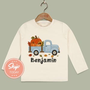 May include: A long-sleeve cream-colored shirt featuring a graphic of a light blue gingham pickup truck carrying orange pumpkins and a sunflower. The name Benjamin is printed in dark gray lettering below the truck illustration.