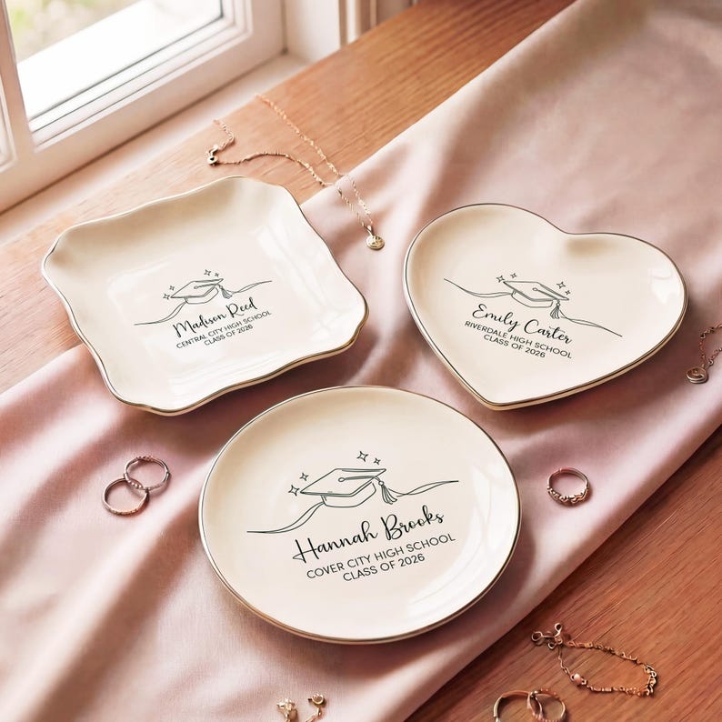 May include: Three ivory-colored ceramic trinket dishes in different shapes: square, heart, and round. Each dish has a gold rim and a graduation cap design with a name and school details. The text reads: Madison Reed, Emily Carter, and Hannah Brooks.