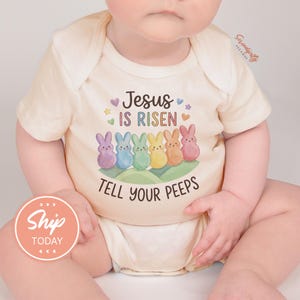 May include: Cream-colored baby bodysuit with the text "Jesus IS RISEN TELL YOUR PEEPS" and a row of colorful bunny illustrations. The design includes purple, blue, green, yellow, and orange bunnies, with hearts and stars.