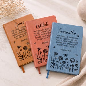 May include: Three rectangular journals in tan, coral, and blue, each featuring a name at the top, the Bible verse Jeremiah 29:11, and black line art of wildflowers and butterflies. Each journal includes a matching ribbon bookmark.