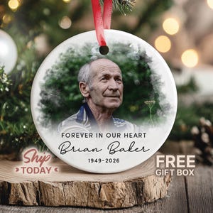 May include: A round white ceramic memorial ornament featuring a portrait of an older man with short hair. The text reads FOREVER IN OUR HEART, Brian Baker, and 1949-2026. A red ribbon is attached to the top for hanging on a tree branch.