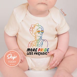 May include: Cream-colored baby onesie with a rainbow graphic of a woman wearing rainbow heart-shaped sunglasses. The text "MORE PRIDE LESS PREJUDICE" is below the graphic. The onesie is on a baby.