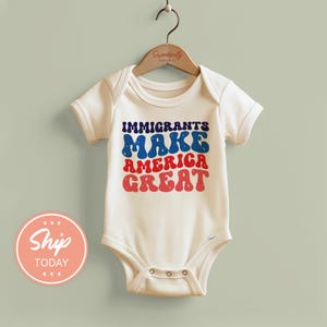 May include: A white baby bodysuit with the text "IMMIGRANTS MAKE AMERICA GREAT" in blue and red retro font. The bodysuit is hanging on a wooden hanger.