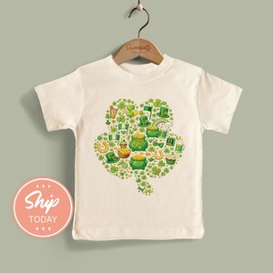 May include: Off-white t-shirt featuring a green shamrock design filled with St. Patrick's Day themed illustrations. The design includes pots of gold, leprechaun hats, and rainbows. The shirt is hanging on a wooden hanger.