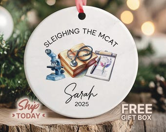 May include: White ceramic Christmas ornament with a red ribbon. The ornament features the text "Sleiging the MCAT" and a watercolor illustration of a microscope, book, stethoscope, pen, and medical chart. The name "Sarah" and the year "2025" are also included.