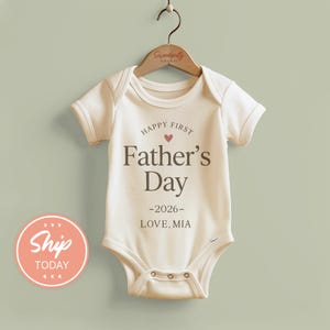 May include: Cream-colored baby onesie with the text "Happy First Father's Day -2026- Love, Mia" in brown lettering. A small red heart is above the words "Father's Day". The onesie is hanging on a wooden hanger.