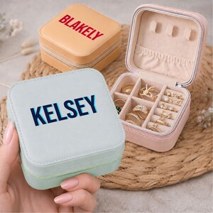 May include: Two square zippered jewelry cases with smooth finishes. One case is light blue with the name KELSEY printed in dark blue, and the other is tan with the name BLAKELY in red. The open case reveals a beige interior with ring slots.