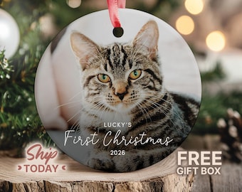 May include: A round white ceramic ornament featuring a photograph of a brown tabby cat with yellow eyes. The text reads Lucky's First Christmas 2026 in white and black lettering. A red ribbon is threaded through a hole at the top for hanging.