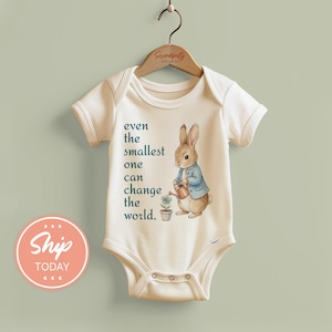 May include: Cream-colored baby bodysuit featuring a watercolor illustration of a rabbit in a blue jacket watering a plant. The text "even the smallest one can change the world" is printed on the front. The bodysuit is hanging on a wooden hanger.