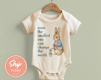May include: Cream-colored baby bodysuit featuring a watercolor illustration of a rabbit in a blue jacket watering a plant. The text "even the smallest one can change the world" is printed on the front. The bodysuit is hanging on a wooden hanger.