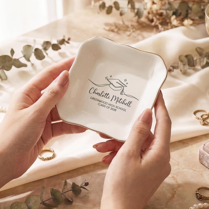 May include: A white ceramic trinket dish with a gold rim, featuring a graduation cap design and the name "Charlotte Mitchell" with "Greenwood High School Class of 2026" printed below. The dish is held by hands.