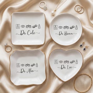 May include: Four white ceramic trinket dishes with gold rims, each featuring an eye chart, eye, glasses, contact lenses, and eye drop graphic. Each dish is personalized with a name: Dr. Cole, Dr. Brown, Dr. Alex, and Dr. Lee.