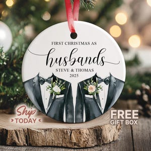 May include: A white, round Christmas ornament with the text "FIRST CHRISTMAS AS husbands" and the names "STEVE & THOMAS 2025". The ornament features illustrations of two tuxedo jackets with floral accents and a red ribbon for hanging.