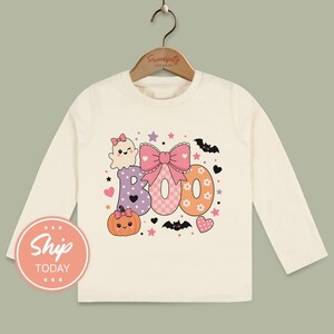 May include: A cream-colored long-sleeve shirt featuring a graphic print of the word BOO in pastel colors. The design includes a smiling ghost, a pumpkin, bats, stars, hearts, and a large pink bow. The shirt hangs on a wooden hanger.