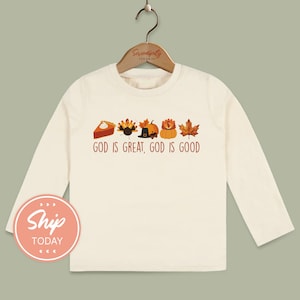 May include: Cream-colored long-sleeved shirt featuring Thanksgiving-themed graphics: a pie slice, turkey, pilgrim hat, pumpkin, and maple leaf. The text "GOD IS GREAT, GOD IS GOOD" is printed below the graphics. The shirt is hanging on a wooden hanger.