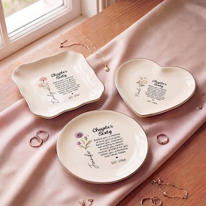 May include: Three cream-colored ceramic trinket dishes in square, heart, and round shapes. Each dish features a floral illustration, the text Chapter Sixty, a name, and EST. 1966. The dishes have gold-toned rims and are arranged on a soft fabric.