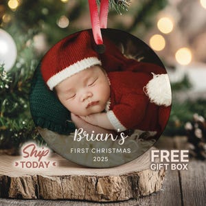 May include: A round Christmas ornament featuring a baby in a Santa Claus outfit. The ornament has the text "Brian's First Christmas 2025". The ornament is hanging from a red ribbon.