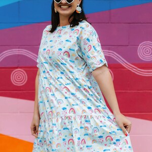 May include: A woman wearing a light blue dress with a rainbow, cloud and sun pattern. She is smiling and wearing heart-shaped sunglasses and colourful earrings. The dress has a ruffled hem.