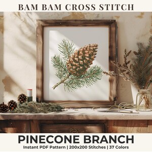 May include: A framed cross stitch pattern titled BAM BAM CROSS STITCH PINECONE BRANCH. The artwork features a detailed illustration of a brown pinecone on a green pine branch against a white background. The text includes 200x200 Stitches and 37 Colors.