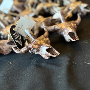 May include: A collection of miniature buffalo skull ornaments, each with intricate detailing. The skulls are painted in shades of brown and gold. A tag with text is attached to one of the skulls. The skulls are displayed on a dark surface.