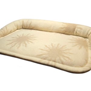 May include: A rectangular pet bed with a raised, cushioned rim. The bed is beige with a sunburst pattern in a lighter shade. The bed is approximately 24 inches long and 18 inches wide.