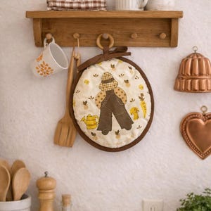 May include: A kitchen scene featuring a wooden shelf with a mug, a small plant, and a checkered cloth. A round, quilted pot holder with a farmer design hangs below. Copper molds and a heart-shaped decoration are also visible.
