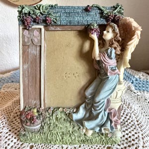 May include: A rectangular picture frame featuring a sculpted angel with wings sitting on a pedestal. The frame includes a wooden door design, a basket of fruit, and grape clusters. The angel wears a blue and pink gown and holds grapes.