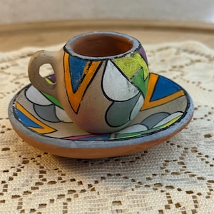 Pode incluir: Um conjunto de chávena e pires em miniatura de terracota com um padrão geométrico pintado à mão em tons vibrantes de laranja, azul, verde, roxo e branco. A superfície de barro mate tem contornos pretos que definem as formas abstratas.