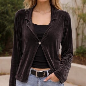 May include: A dark gray velvet blazer with a single button closure. The blazer has long sleeves and a notched collar. The button is a round, silver-toned jewel. The person is wearing a black top and blue jeans.