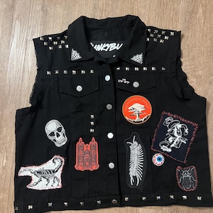 May include: Black denim vest with silver studs and various patches, including a skull, a scorpion, a building, a centipede, a mushroom, an eye, and a spider. The vest has a collar with a spiderweb design and the text "INKYBU" on the back.