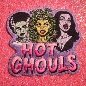 May include: An embroidered patch featuring three female figures with green, yellow, and pale skin tones, set against a purple background. The text HOT GHOULS is stitched in pink and white gradient lettering at the bottom of the patch design.