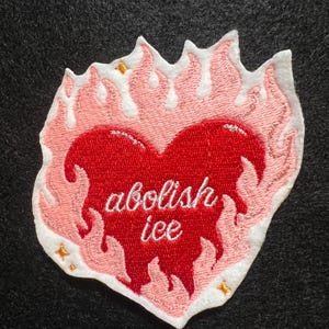 May include: Embroidered patch featuring a red heart with the words "abolish ice" in white script. The heart is surrounded by pink flames with white outlines and small gold star accents. The patch is on a dark gray background.