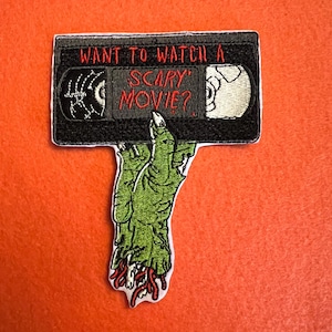 Op de afbeelding: Geborduurde patch met een zwarte VHS-band met de tekst "WANT TO WATCH A SCARY MOVIE?" en een groene zombiehand. De patch heeft een witte rand en is geplaatst tegen een oranje achtergrond.