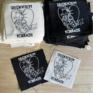 May include: A collection of square fabric patches in black and off-white. Each patch features a screen-printed graphic of a baby with a mohawk inside a heart, accompanied by the text PUNKYBUTT THREADS. The patches are arranged in two stacks.