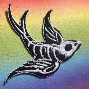 Op de afbeelding: Zwart en wit geborduurde patch van een vogel in vlucht, met een skeletontwerp. De patch heeft een gedetailleerd ontwerp met witte stiksels op een zwarte achtergrond. De achtergrond is een regenboogverloop.