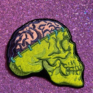 May include: Embroidered patch featuring a green skull with a pink brain and blue stitching. The skull has a stitched appearance, with a black border. The patch is set against a sparkly purple background.