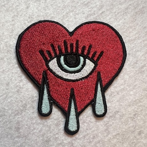 May include: Embroidered patch in the shape of a red heart with a white and black eye in the centre. The heart has three blue teardrops. The patch is on a grey background.