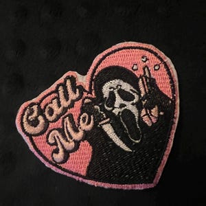 May include: Heart-shaped embroidered patch with a pink background and black trim. The patch features the Ghostface mask from the Scream films, holding a knife. The words "Call Me" are embroidered in pink.