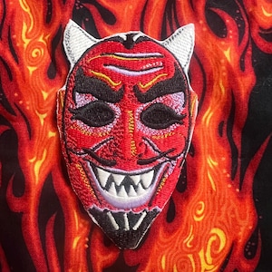 May include: An embroidered patch featuring a red devil mask with white horns, black eyebrows, and sharp white teeth. The mask has detailed stitching in red, black, and white thread, showcasing a pointed chin and a menacing expression on a fabric background.