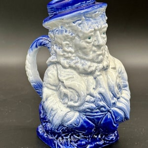 Puede incluir: Una jarra de cerámica azul y blanca con forma de hombre con barba y sombrero. La jarra tiene un asa y un pico.