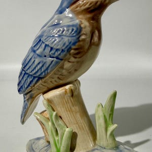 Könnte beinhalten: Eine Keramikfigur eines Eisvogels, der auf einem Baumstumpf sitzt. Der Vogel hat einen glänzenden blauen Kopf und Flügel, eine hellbraune Brust und einen langen, spitzen Schnabel. Der Sockel ist mit grünem Gras und blauem Wasser bemalt.