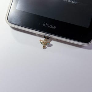 May include: A Kindle e-reader with a clear protective case. A gold-colored charm in the shape of an ankh is attached to the bottom of the device. The Kindle screen displays text and star ratings.