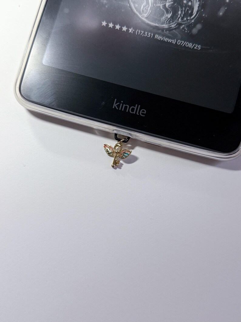 May include: A Kindle e-reader with a clear protective case. A gold-colored charm in the shape of an ankh is attached to the bottom of the device. The Kindle screen displays text and star ratings.