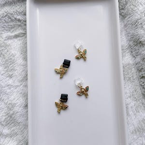 May include: Four small gold-colored angel wing charms with colorful enamel details and black clips are displayed on a white rectangular tray. The charms have a small clear plastic piece attached.