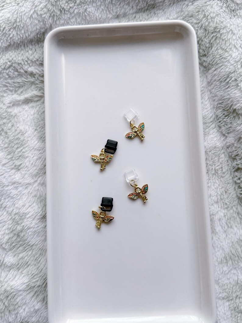 May include: Four small gold-colored angel wing charms with colorful enamel details and black clips are displayed on a white rectangular tray. The charms have a small clear plastic piece attached.