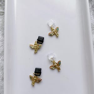 May include: Four small, gold-colored charms with a key and wing design. The wings are multi-colored. Two charms have black clips, and two have clear plastic attachments. The charms are on a white surface.
