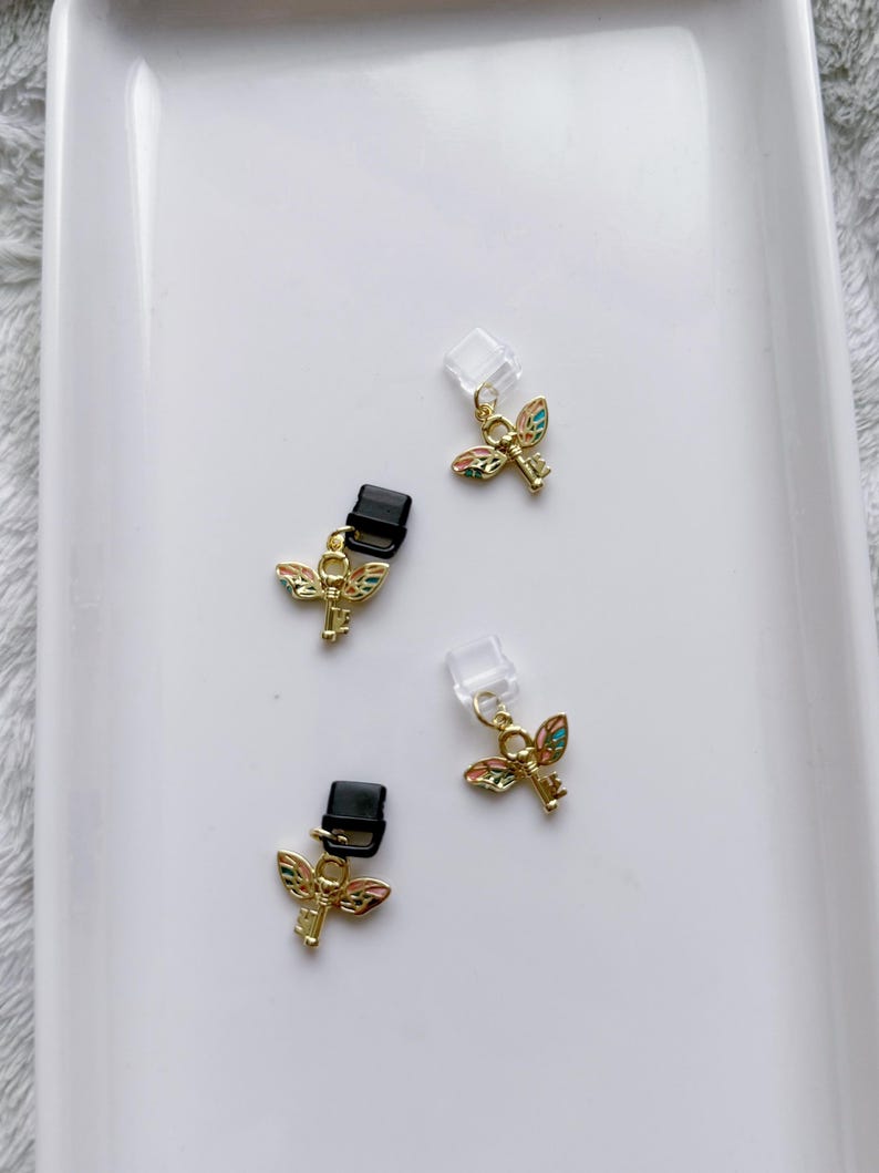 May include: Four small, gold-colored charms with a key and wing design. The wings are multi-colored. Two charms have black clips, and two have clear plastic attachments. The charms are on a white surface.