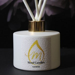 May include: White diffuser bottle with a gold lid and sticks. The bottle has a label with a gold drop and the text "Mood Candles Gardenia".